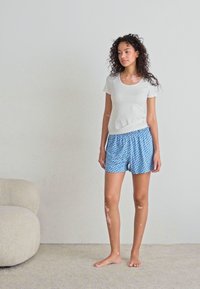 Ljust grå kortärmad skjorta och blå mönstrade shorts med elastisk midja. Modellen står barfota på ett strukturerat golv nära en soffa.