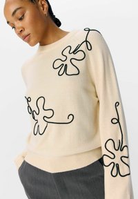 Pullover crema con ricami floreali neri sul davanti e sulle maniche, caratterizzato da un colletto e orlo a coste. Texture morbida e vestibilità ampia.