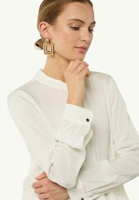 Weiße Bluse mit langen Ärmeln aus weichem Stoff, ausgestattet mit einem Stehkragen und schwarzen Knopfdetails an den Manschetten. Das Modell trägt große goldene Ohrringe.