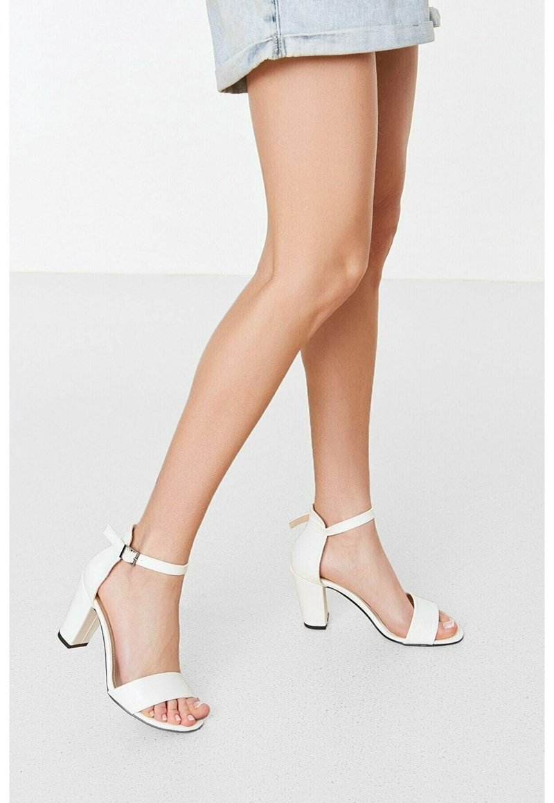 Jambes portant un short en jean clair et des sandales blanches à talons hauts avec brides autour de la cheville, sur un sol clair et un fond blanc.