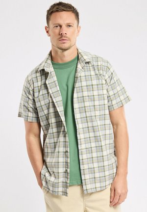 Homme portant un t-shirt vert sous une chemise à carreaux à manches courtes ouverte, les mains dans les poches d'un pantalon beige sur fond blanc.