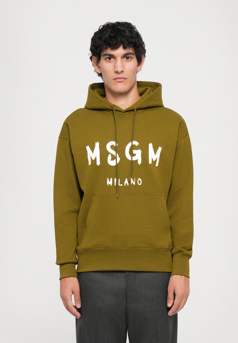 Jaunas vyras, vilkintis alyvų žalios spalvos gobtuvu su "MSGM MILANO" tekstu, stovintis prieš paprastą baltą foną.