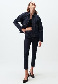 Giacca di denim scuro con taglio cropped, due tasche frontali e cuciture in evidenza; abbinata a pantaloni skinny di denim scuro e tacchi neri.