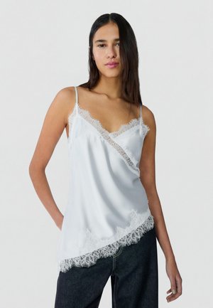 Vrouw die een wit satijnen hemdje met kanten rand en donkere spijkerbroek draagt, staand tegen een effen lichte achtergrond.