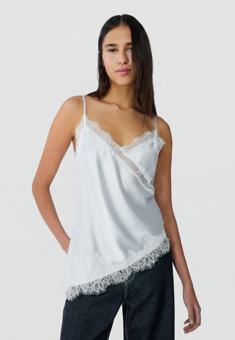 Femme portant une camisole en satin blanc avec bordure en dentelle et un jean foncé, debout devant un fond uni clair.
