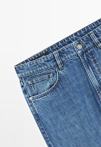 Jean en denim bleu avec un design traditionnel à cinq poches, comprenant un bouton métallique visible et des coutures contrastées. La texture est douce avec un léger délavage.