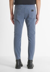 Hombre con pantalones ajustados azules y botas negras hasta el tobillo, de pie contra un fondo claro, visto desde atrás.