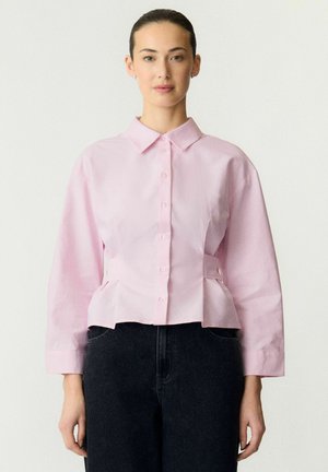 Kvinde iført en lyserød bluse med knapper og lange ærmer samt mørke, højtaljede jeans, stående mod en ensfarvet lys baggrund.