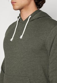Mikina s kapucí v tmavě zelené barvě, vyrobená z měkké látky. Má klokaní kapsu a kontrastní bílé šňůrky na kapuci.