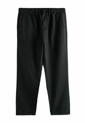 Férfi fekete zsinóros nadrágot és fehér inget visel, mutatja a derékrészt és a nadrág mellett pihenő kezét.