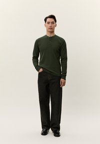 Férfi áll sötétzöld, hosszú ujjú henley ingben, fekete bő, lazán eső farmernadrágban és fekete cipőben, egyszínű világos háttér előtt.