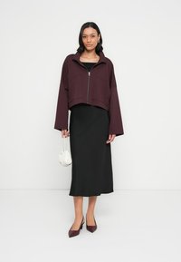 Krótka bordowa kurtka z wysokim kołnierzem i przednim zamkiem błyskawicznym, zestawiona z długą czarną dopasowaną spódnicą i bordowymi butami na obcasie, trzymająca białą torbę.