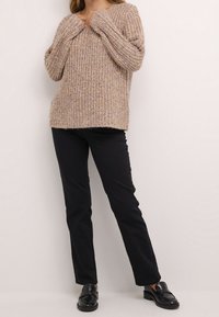 Pull en tricot beige à coupe décontractée et surface texturée, associé à un pantalon noir à jambe droite et des mocassins noirs.