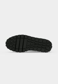 Semelle de chaussure en caoutchouc noir avec des motifs de bande de roulement profonds et multidirectionnels pour la traction, présentée sur un fond blanc uni.