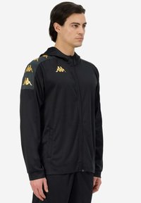 Giacca sportiva nera con zip e cappuccio, con loghi Kappa dorati sulle spalle, realizzata in materiale liscio e leggero.