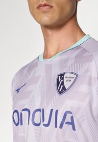 Gros plan sur une personne portant un maillot de football VfL Bochum violet clair avec des motifs géométriques et un texte de sponsor en bleu.