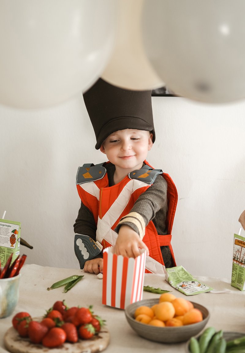 Kind draagt een oranje-grijze outfit met een hoge zwarte hoed, reikt in een gestreepte popcorndoos, omringd door fruit en groenten.