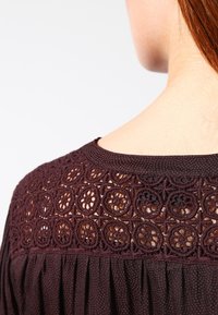 Zbliżenie na górną część pleców kobiety w ciemno-brązowej bluzce z okrągłym wzorem koronkowym w kwiaty na karczku.