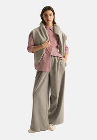 Jasnoszara wełniana kamizelka nałożona na pasiasty czerwono-biały t-shirt, w połączeniu z szerokimi spodniami w szary prążek. Model nosi białe buty.