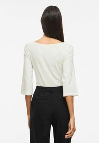 Sieviete ar gariem tumšiem matiem, tērpusies baltā trīs ceturtdaļu piedurkņu topā un melnās augsta vidukļa biksēs, redzama no aizmugures uz balta fona.