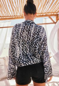 Schwarze und weiße gemusterte transparente Bluse mit langen Ärmeln, kombiniert mit dunklen Shorts. Der Hintergrund zeigt eine Holzstruktur und sanfte Stoffvorhänge.