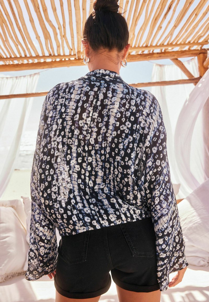 Schwarze und weiße gemusterte transparente Bluse mit langen Ärmeln, kombiniert mit dunklen Shorts. Der Hintergrund zeigt eine Holzstruktur und sanfte Stoffvorhänge.