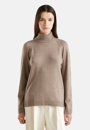 Femme debout, portant un pull col roulé beige clair et un pantalon couleur crème, sur un fond blanc uni.