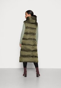 Gilet lungo imbottito di colore verde oliva con cappuccio, design a righe verticali e spacchi laterali, indossato sopra un top verde chiaro e leggings neri.