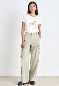 White short-sleeve t-shirt with a brown dog print, paired with light beige cargo pants featuring side pockets and zipper accents, worn with black shoes.