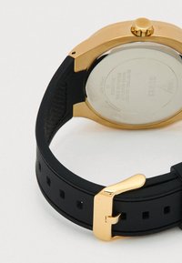 Rückansicht einer Armbanduhr in Goldoptik mit schwarzem Silikonarmband und goldener Schnalle, zeigt die Rückseite des Gehäuses aus Edelstahl mit eingravierten Details.