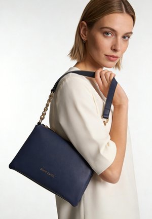 Femme aux cheveux blonds courts portant un haut blanc tient un sac à main Pierre Cardin bleu marine avec une bandoulière chaîne dorée sur l'épaule.