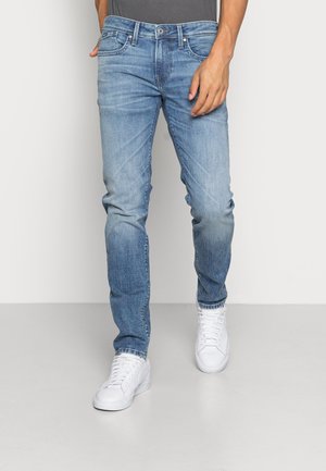 Mann trägt hellblaue ausgewaschene Jeans und weiße Sneaker, steht vor weißem Hintergrund mit einer Hand in der Nähe der Tasche.