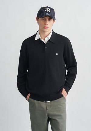 Giovane uomo che indossa una polo nera a maniche lunghe, una camicia bianca sotto, pantaloni color oliva e un berretto da baseball blu navy a righe con un logo bianco.