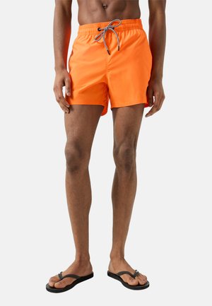 Mężczyzna w jasnopomarańczowych kąpielówkach z czarno-białym sznurkiem i czarnych klapkach, stojący na jednolitym tle.