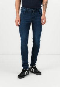 Slim-fit niebieskie dżinsy z ciemnym praniem, z pięcioma kieszeniami, paskiem oraz zapięciem na guziki. Noszone z czarnymi trampkami.