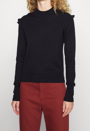 Femme portant un pull noir à manches longues avec des détails à volants aux épaules et un pantalon taille haute couleur rouille sur un fond uni.