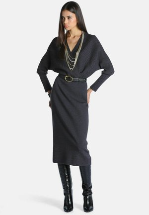 Femme portant une robe en maille gris foncé ceinturée avec de larges manches, des colliers chaînes superposés et des bottes noires montantes, debout les mains sur les hanches.