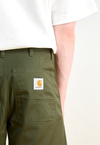 Pantaloni verzi măslinii cu o textură netedă, având un eticheta albă proeminentă cu un logo galben pe buzunarul din spate.