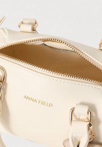 Crèmekleurige handtas met gouden accenten en het reliëflogo "ANNA FIELD". Voorzien van een ritssluiting en textuuraccenten op het handvat.
