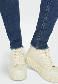 Zapatillas deportivas blancas de lona con una suela texturizada y elevada. Llevadas con unos jeans ajustados azul oscuro, que presentan dobladillos deshilachados en el tobillo.