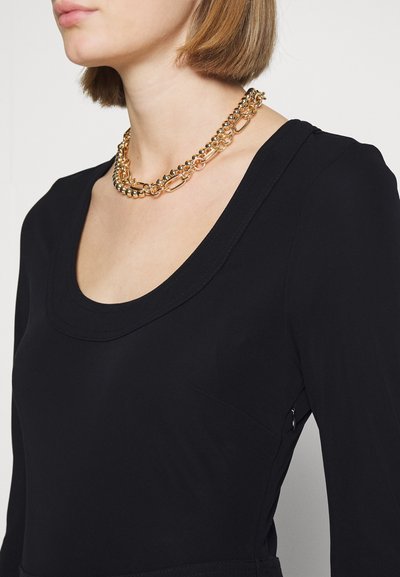 Collier en or avec des maillons ovales et des accents en perles porté sur une robe noire à manches longues avec un col rond et une texture lisse.