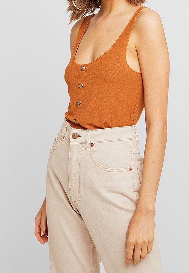 Femme portant un haut orange sans manches boutonné et un pantalon beige taille haute, montrée des épaules aux hanches sur un fond uni.