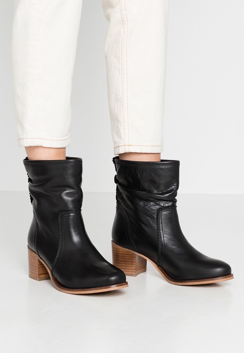 Bottes chevilles en cuir noir à design décontracté, avec un talon bloc en bois et des détails cousus. Portées avec un pantalon clair retroussé.