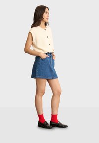 Krämfärgad ribbad kortärmad topp med bruna knappar, i kombination med en blå denimkjol och svarta loafer-skor. Röda strumpor syns.