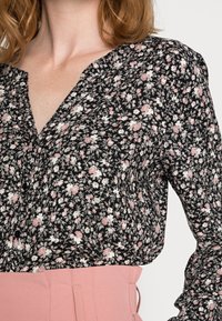 Blouse noire à motif floral avec des motifs roses et blancs, décolleté en V, détails de boutons, et en tissu doux, associée à une jupe taille haute rose clair.
