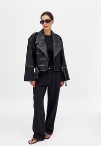 Must must nahkakate, millel on kontrastsed valged õmblused, laiad varrukad ja nööbid, koos vabade mustade pükste ja sandaalidega.