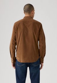 Bruine corduroy shirt met een puntige yokedesign. Beschikt over lange mouwen en met knopen sluitingen. Gecombineerd met donkere denim jeans.