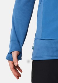Blaues Langarmhemd aus strukturiertem Stoff, mit verstärkten Bündchen und einer Nahtdetails, versehen mit einem kleinen gelben und blauen Etikett am Saum.