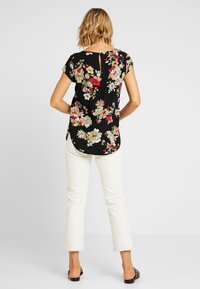 Blommig svart blus med korta ärmar, rund hals och en nyckelhålsdetalj i ryggen. Matchad med vita avslappnade byxor och svarta sandaler.