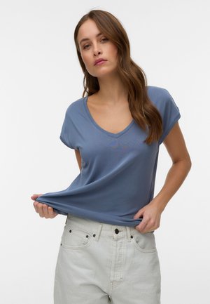 Moteris su ilgais rudais plaukais, dėvinti mėlyną V formos iškirptės marškinėlį ir šviesiai pilkas kelnes, šiek tiek tempia marškinėlių apačią į šoną.